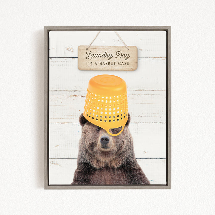 A framed image of a bear wearing a yellow plastic basket on its head, with a wooden sign above it that says "Laundry Day I'm a basket case".