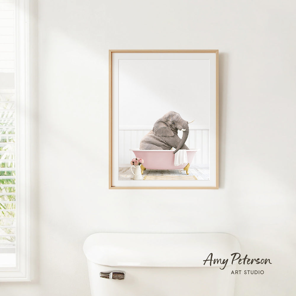 A framed image of a gray elephant sitting in a pink bathtub, with a vase of flowers nearby, is hanging on a white wall.
