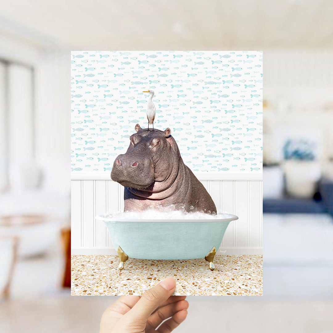 A hand holds up a framed image depicting a hippopotamus sitting in a bathtub, with a seagull perched on its head.