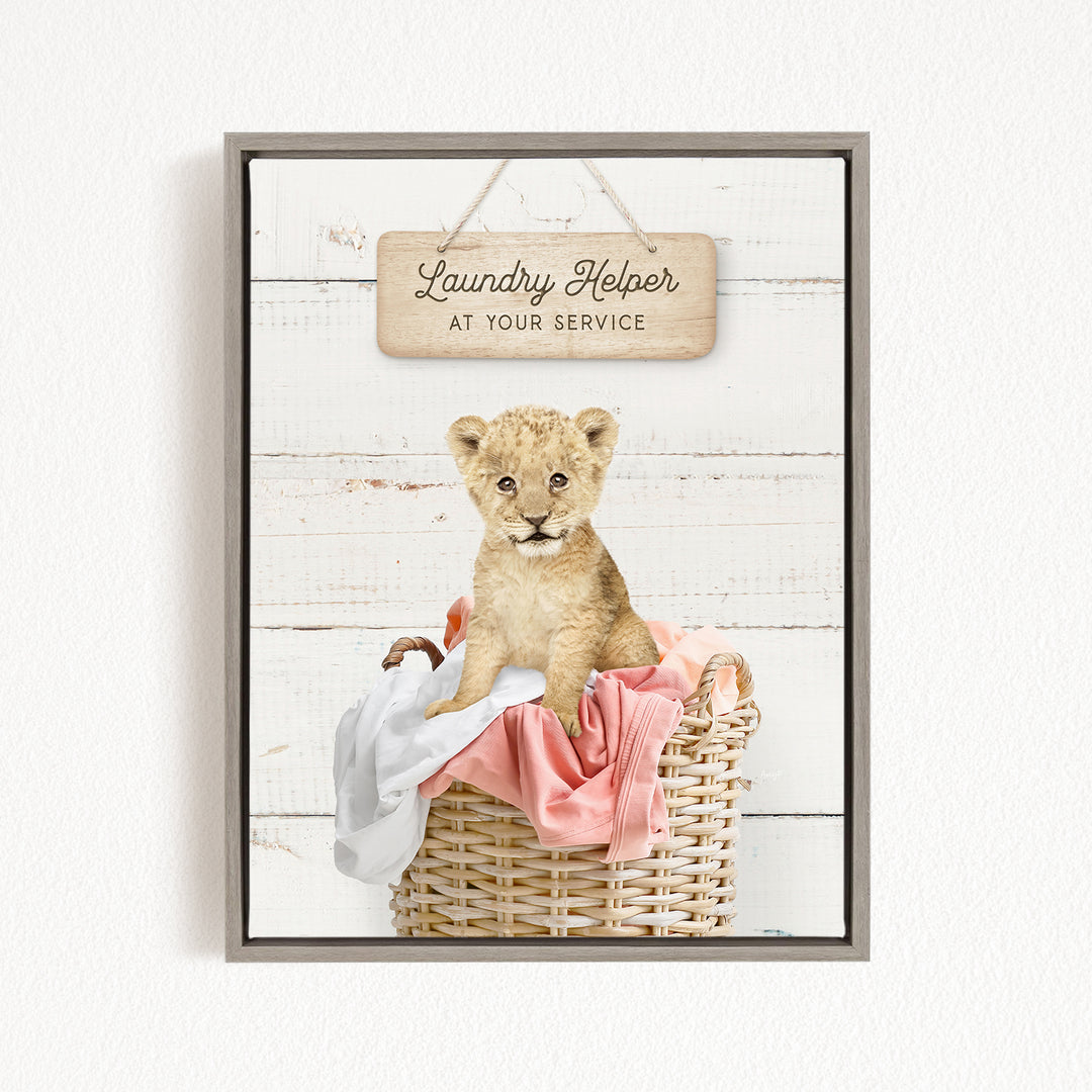 A young lion cub sitting in a wicker basket filled with clothes, with a wooden sign above that reads "Laundry Helper at your service".