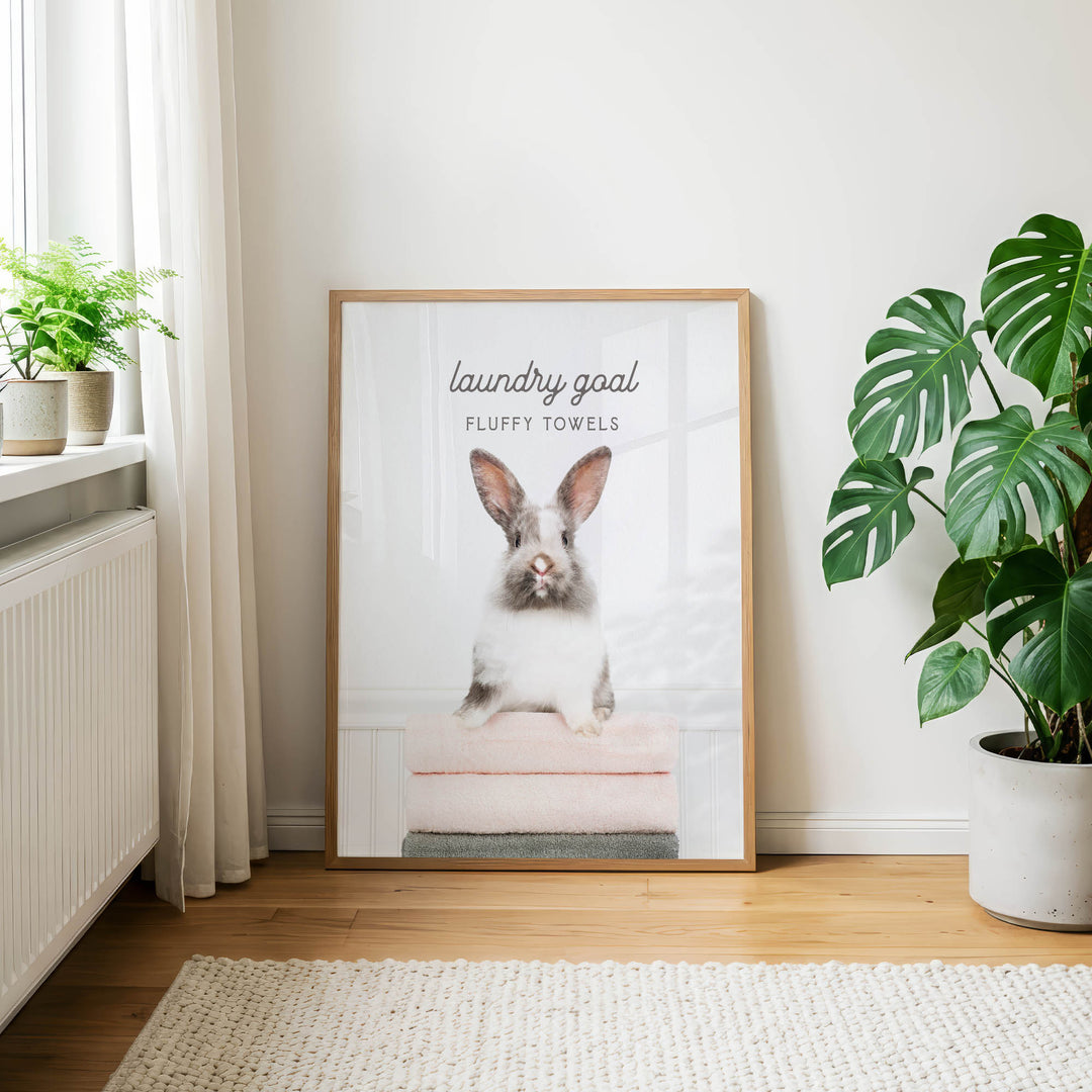 A framed poster of a rabbit sitting on a stack of fluffy towels, with the text "laundry goal" and "FLUFFY TOWELS" printed on it, is displayed on a wall in a room with a window and potted plants.