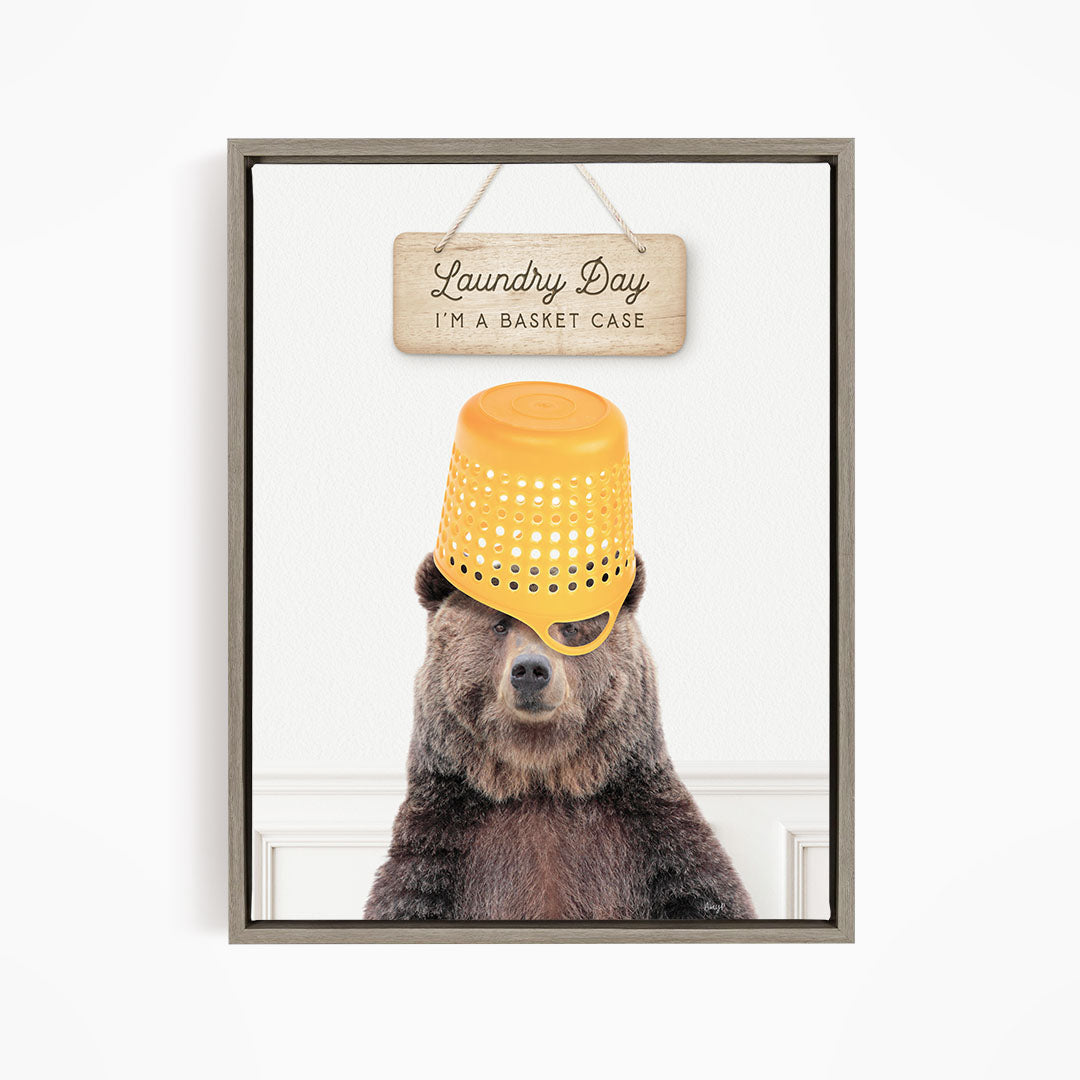 A framed image of a bear wearing a yellow plastic basket hat, with a wooden sign above it that says "Laundry Day I'm a basket case".