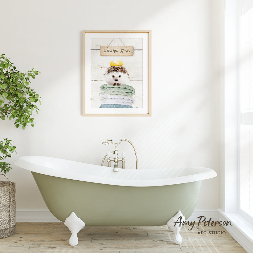 A framed print of a hedgehog sitting on a stack of towels hangs on a white wall above a green clawfoot bathtub in a bathroom.
