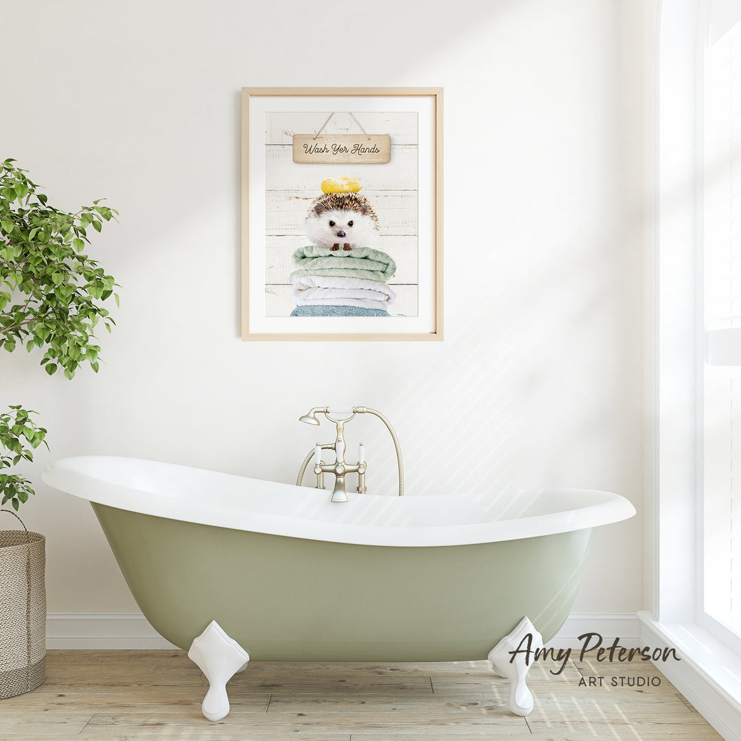 A framed print of a hedgehog sitting on a stack of towels hangs on a white wall above a green clawfoot bathtub in a bathroom.