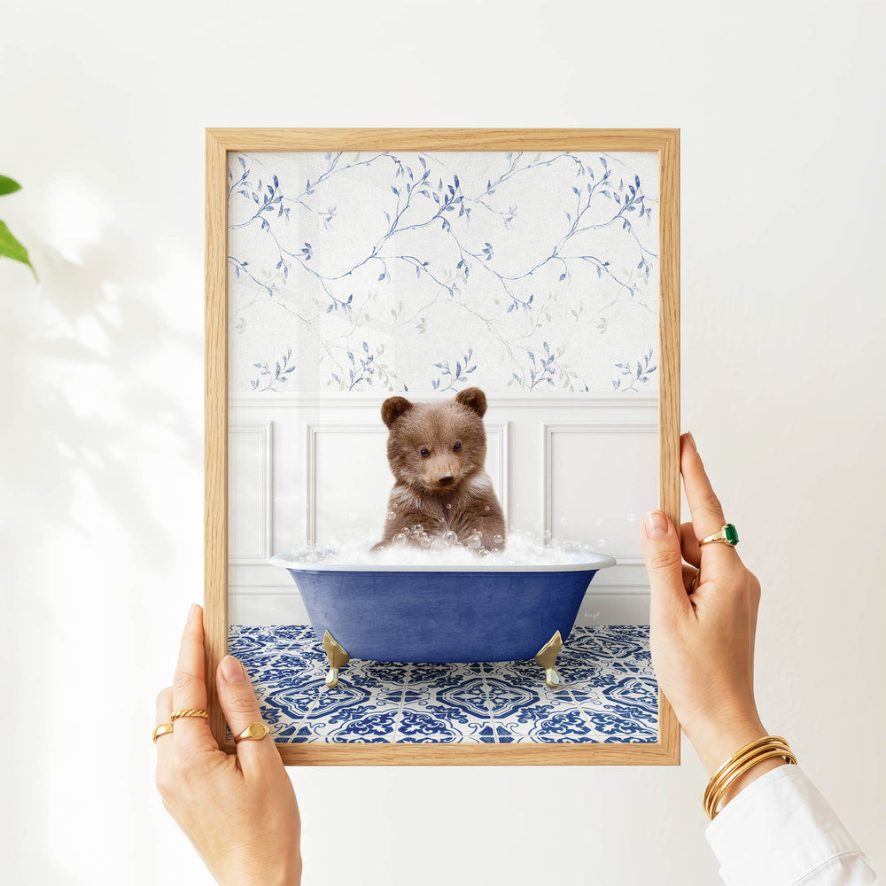 A person's hands are holding a framed picture of a small brown bear sitting in a blue bathtub filled with water, surrounded by a blue and white floral pattern.