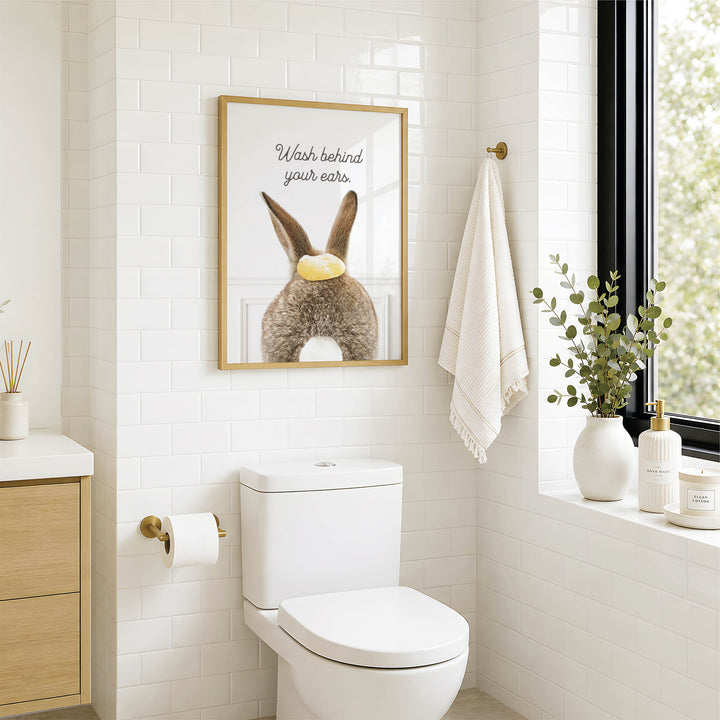A bathroom with a white toilet, a framed poster of a rabbit with a yellow object on its ear, and a window with greenery on the sill.