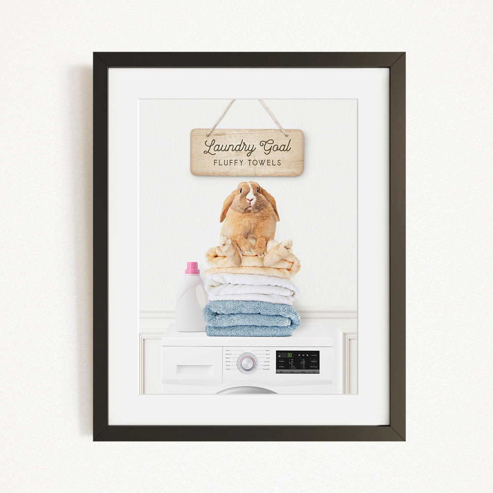 A framed image of a rabbit sitting on a stack of fluffy towels, with a sign above that says "Laundry goal Fluffy towels".