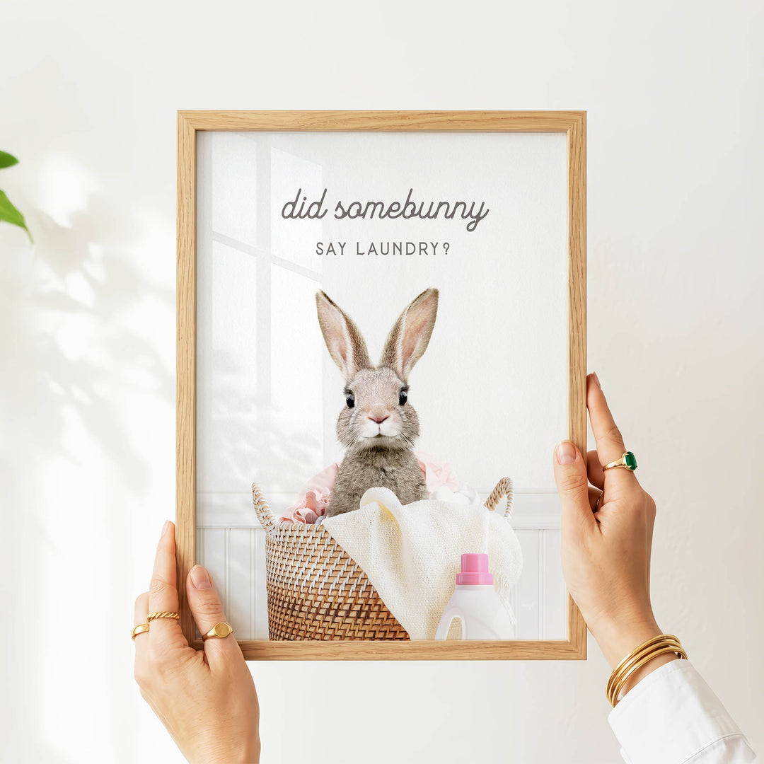 A person is holding up a framed picture of a rabbit sitting in a basket, with the text "did some bunny say laundry?" displayed on the picture.