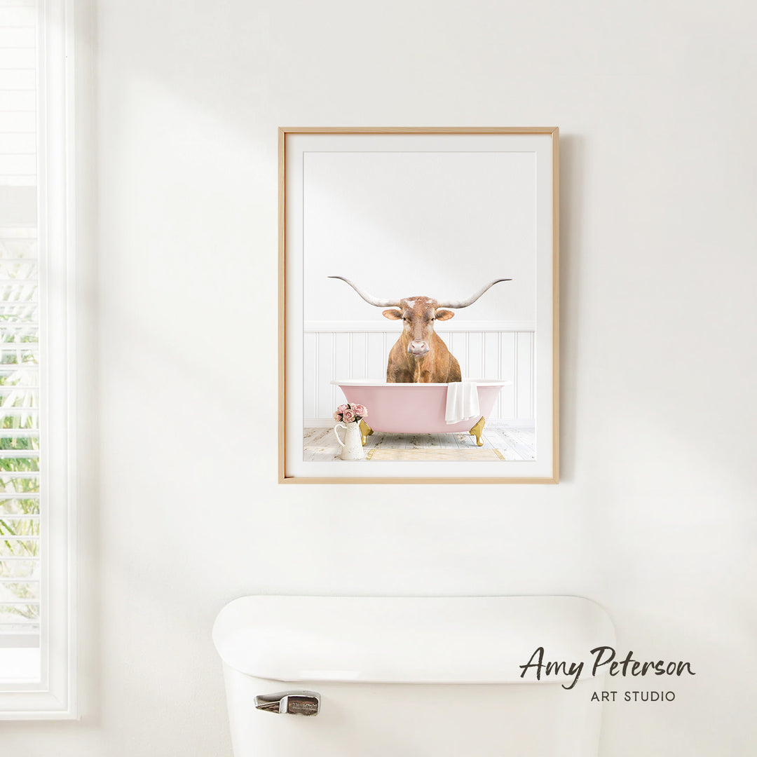 A framed image of a brown bull with long horns sitting in a pink bathtub, with a vase of flowers and a white toilet in the foreground.
