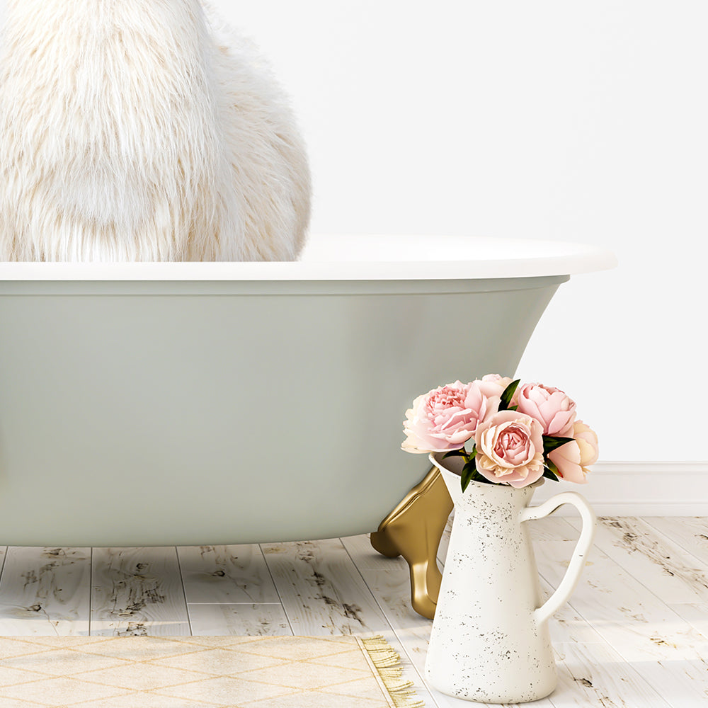 A white cat is sitting in a green bathtub, with a vase of pink flowers placed next to it on a wooden floor.