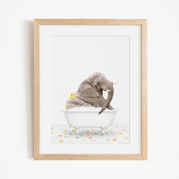 A gray elephant is sitting in a white bathtub filled with bubbles, with a yellow rubber duck floating on the water.