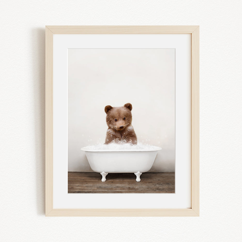 A cute brown bear sitting in a white bathtub filled with bubbles, looking directly at the camera.