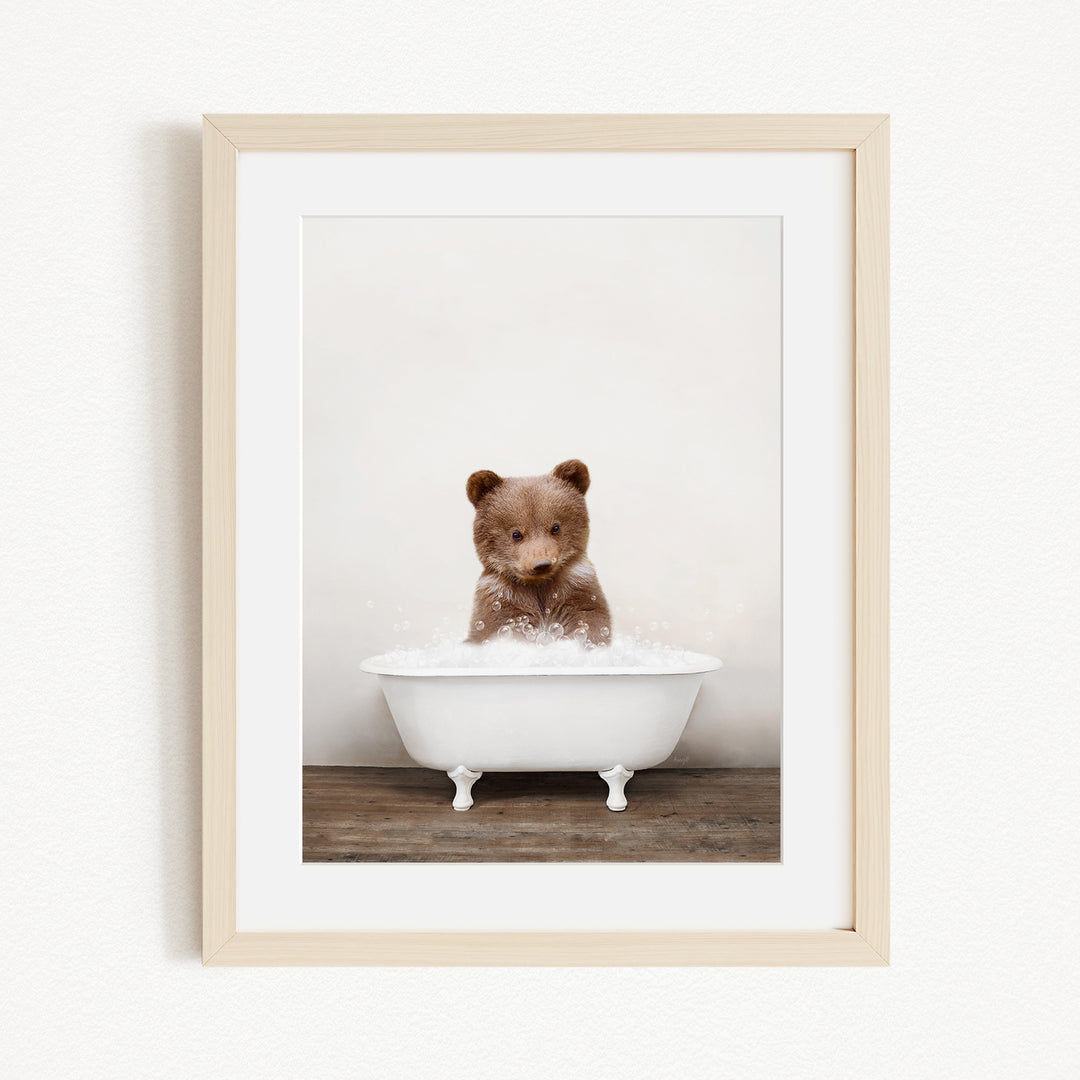 A cute brown bear sitting in a white bathtub filled with bubbles, looking directly at the camera.