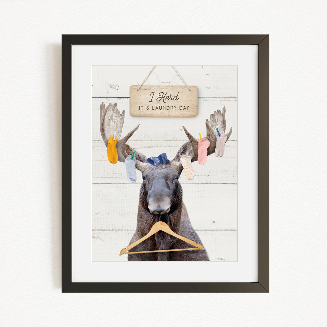 A framed image depicting a moose with a sign that says "I herd, it's laundry day" hanging on a wooden background.