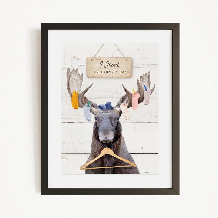 A framed image depicting a moose with a sign that says "I herd, it's laundry day" hanging on a wooden background.