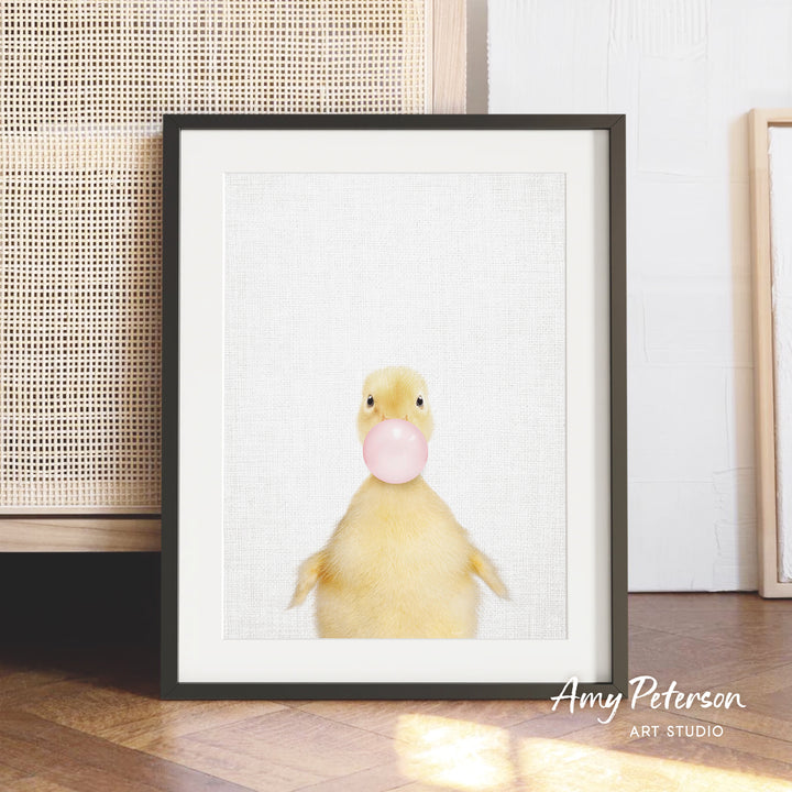 A framed image of a yellow duck blowing a bubble with a pink bubblegum bubble.