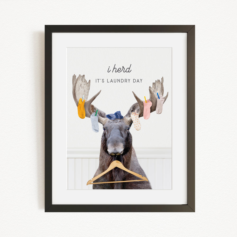 A framed image depicting a moose with a laundry basket hanging from its antlers, accompanied by the text "It's laundry day"
