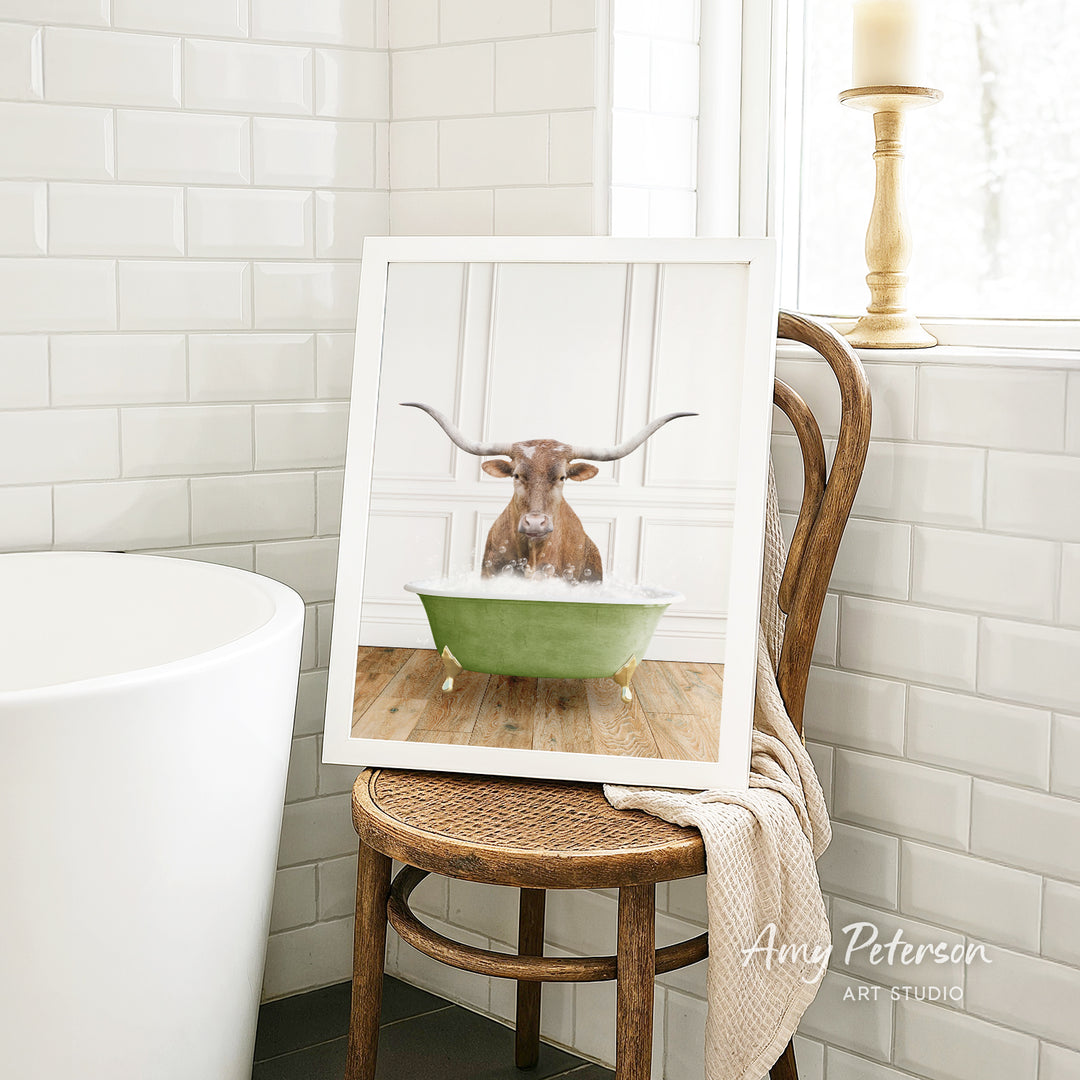 A framed image of a bull with long horns sitting in a green bathtub, surrounded by white tiles and a wooden chair.