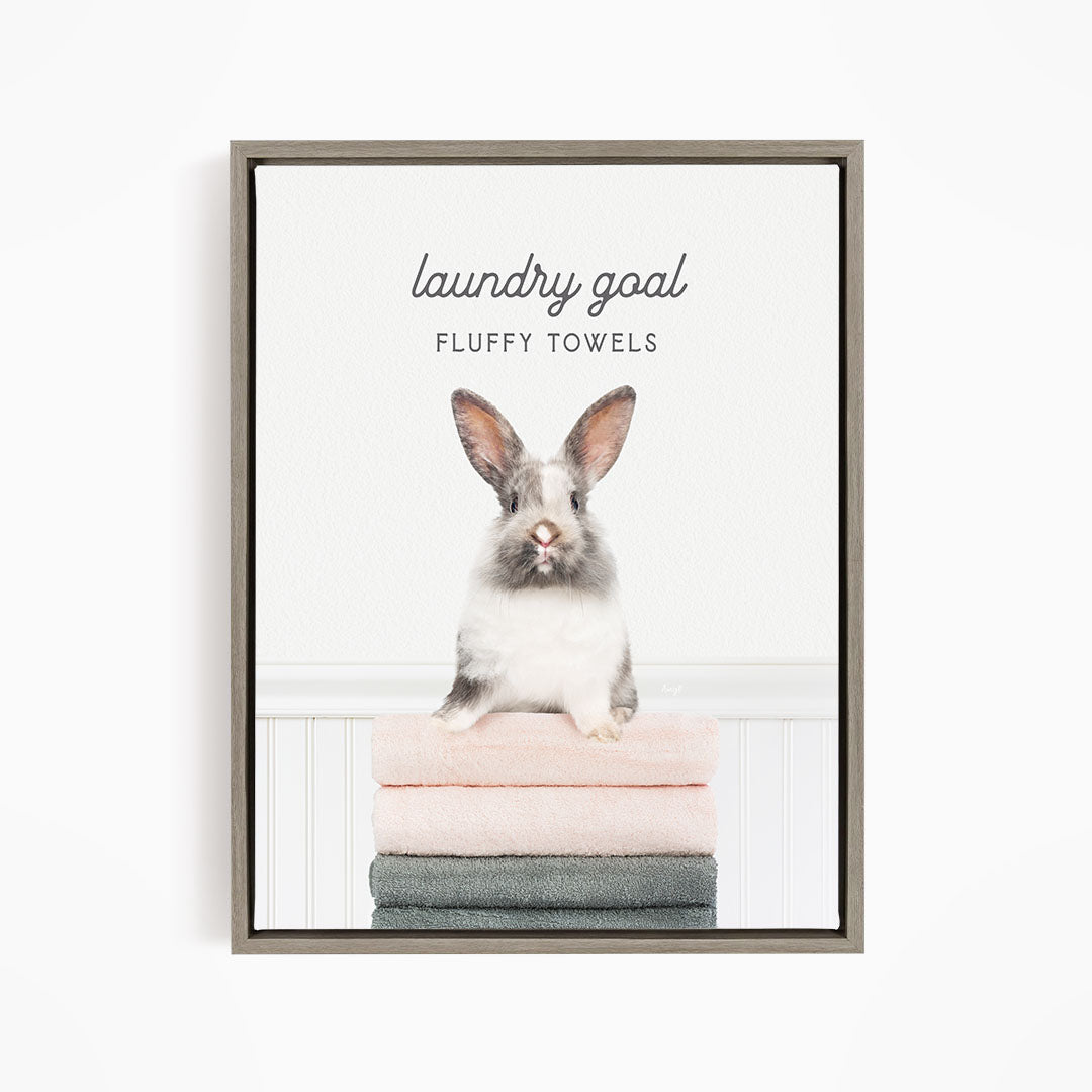 A fluffy white rabbit sitting on a stack of towels, with the text "laundry goal FLUFFY TOWELS" displayed above the rabbit.