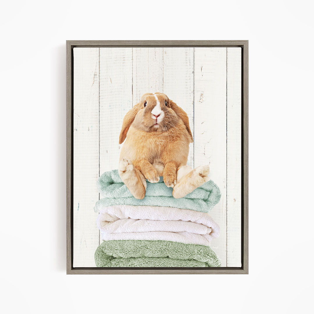 A brown rabbit sitting on a stack of green and white towels against a white wooden background.