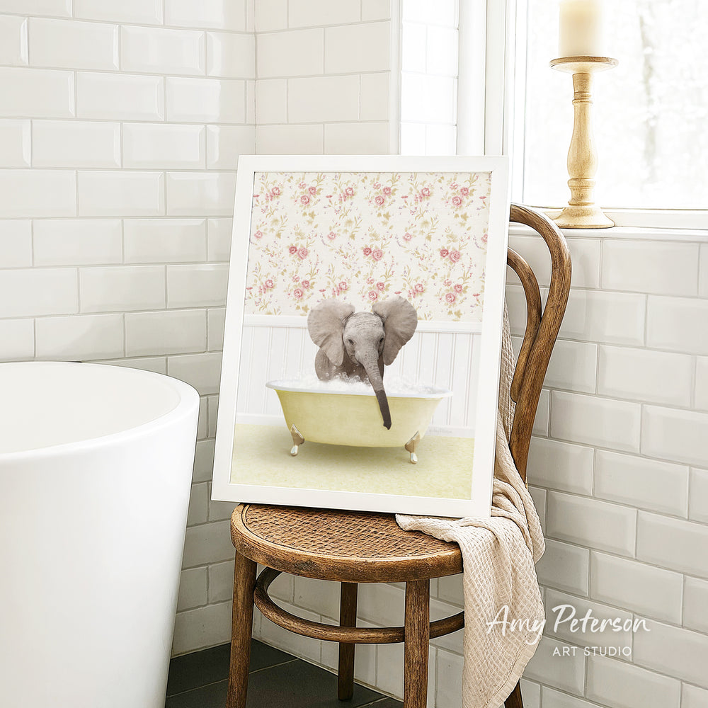 A framed image of a baby elephant sitting in a bathtub, surrounded by a floral pattern, is displayed on a wooden chair in a bathroom setting.
