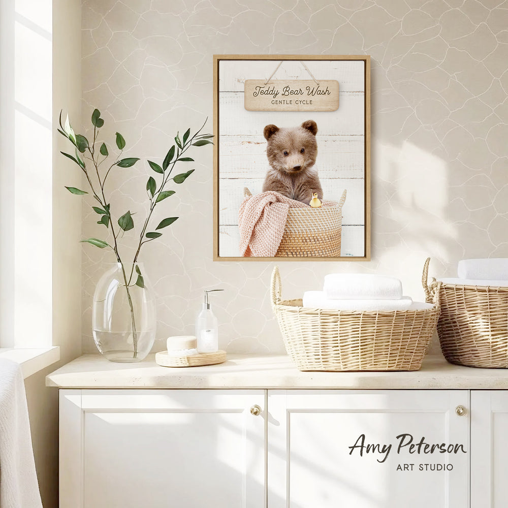 a framed photo of a teddy bear sitting in a basket, with a sign above it that reads "Teddy Bear Wash Gentle Cycle". The photo is displayed on a wall above a white cabinet, which has a vase of leaves and a bottle of soap on it.