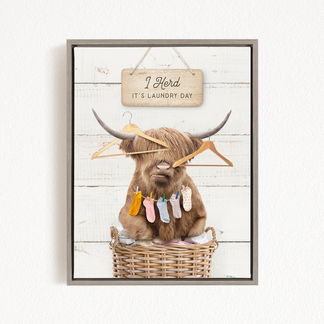A framed image depicting a Highland cow sitting in a wicker basket, surrounded by clothes and hangers, with a sign above that reads "I herd it's laundry day".