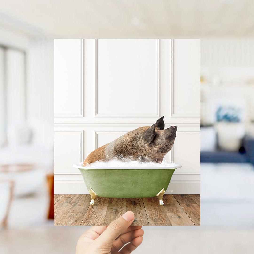 A hand holds up a framed image of a pig sitting in a green bathtub filled with bubbles, with a blurred background suggesting a bathroom setting.