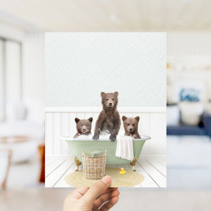 A hand holds up a framed image depicting three adorable brown bears sitting in a green bathtub, surrounded by a yellow rubber duck and a white towel.