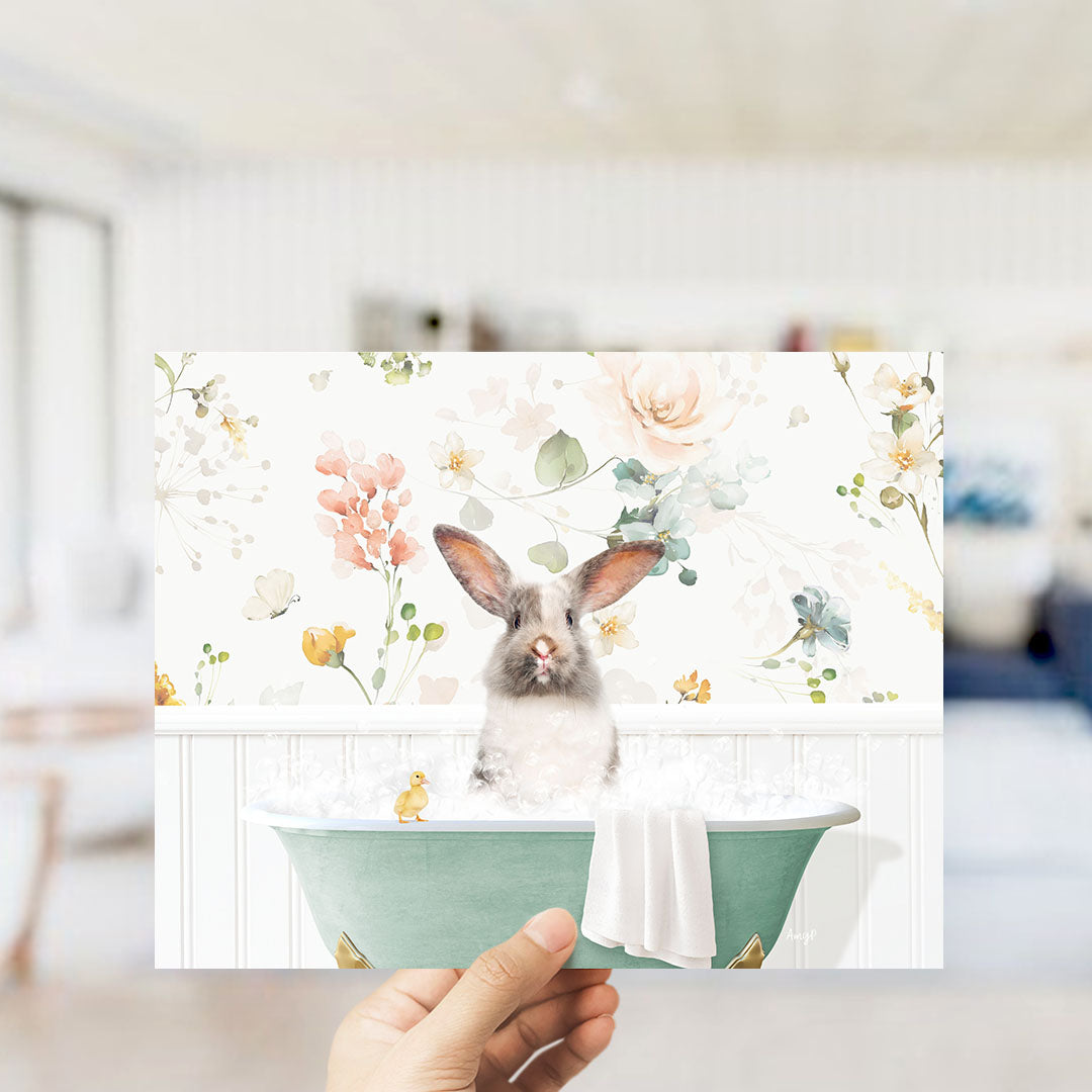 A hand holding a framed print featuring a cute rabbit sitting in a bathtub filled with water and surrounded by flowers.