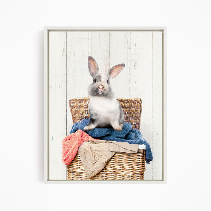 A gray and white rabbit sitting in a wicker basket filled with clothes and towels against a white wooden background.