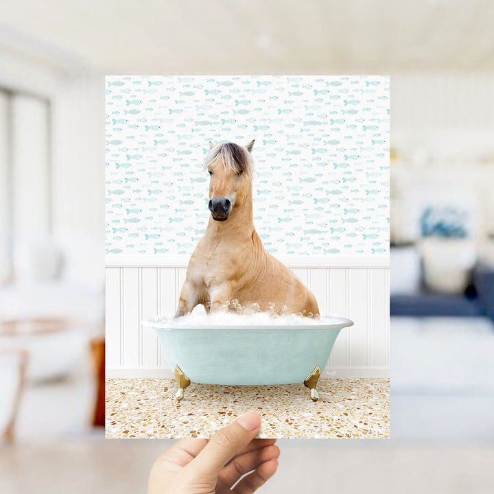 A hand holds up a framed image of a horse sitting in a bathtub filled with bubbles, surrounded by a pattern of small fish.