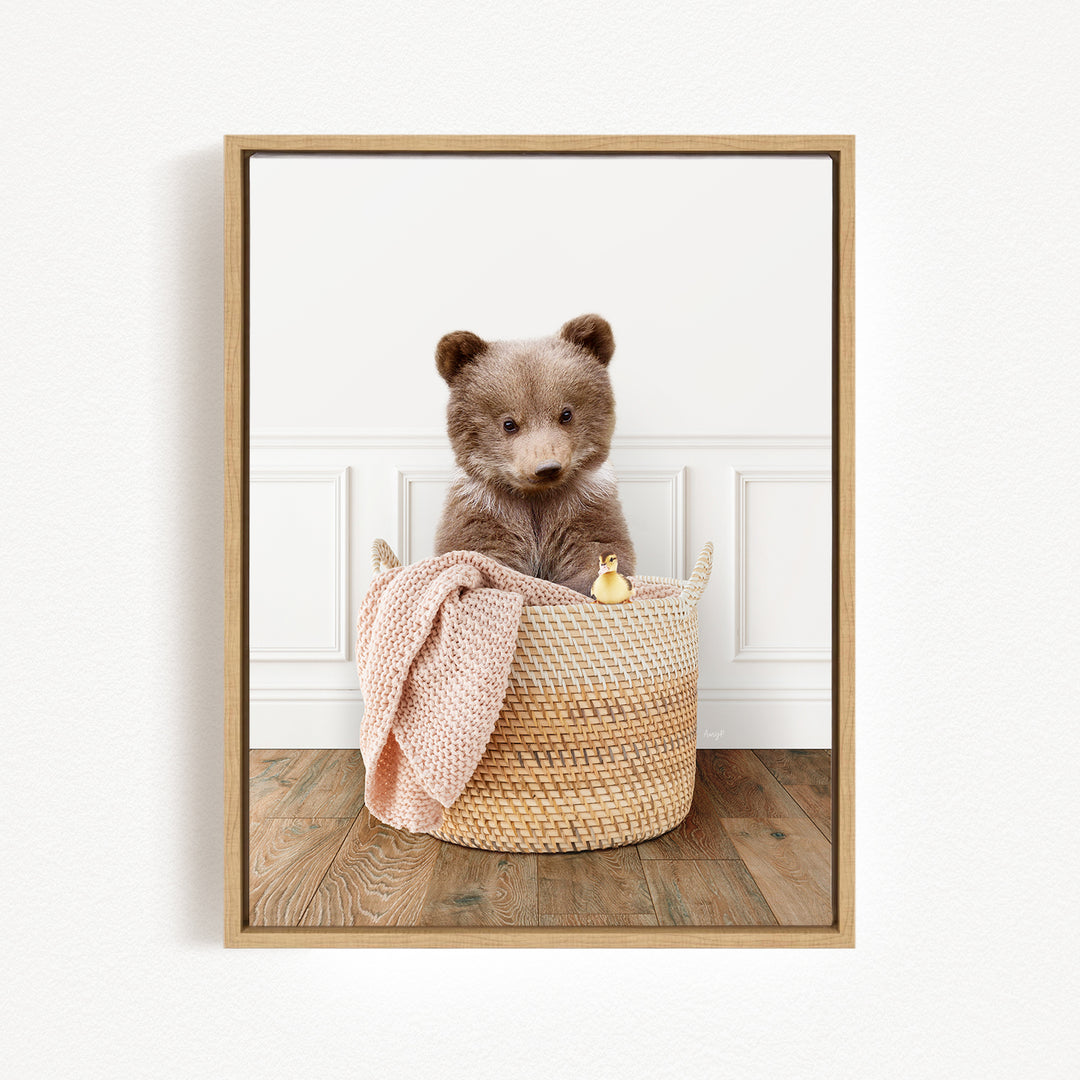 A brown bear sitting in a woven basket, with a pink blanket and a small yellow toy duck nearby.