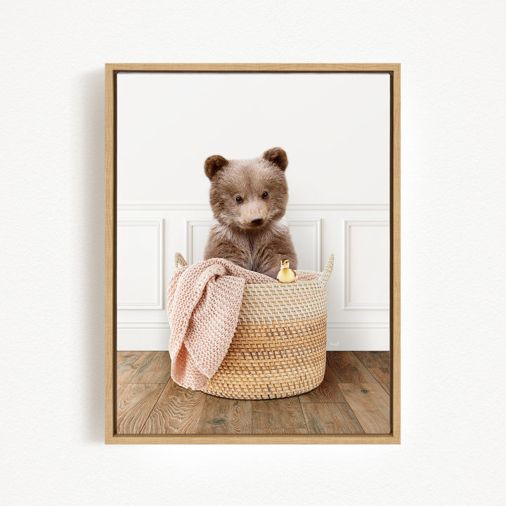 A brown bear sitting in a woven basket, with a pink blanket and a small yellow toy duck nearby.