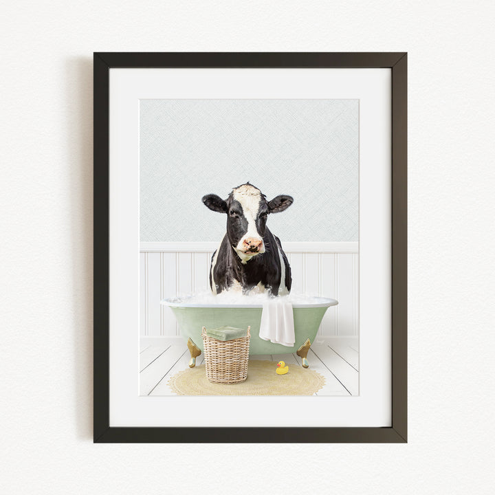 A black and white cow is sitting in a green bathtub filled with water, surrounded by a basket of rubber ducks and a yellow rubber ducky.