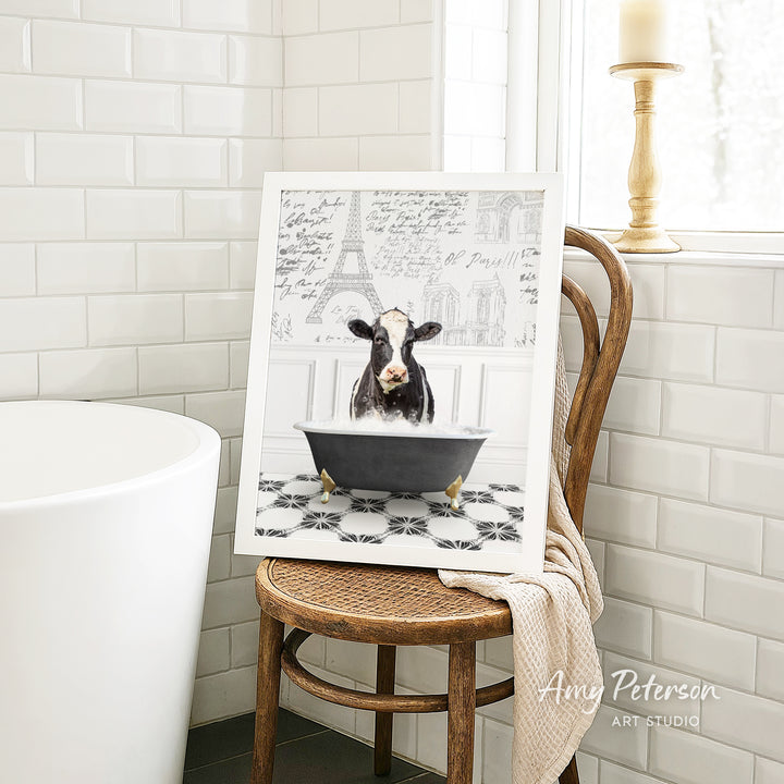 A framed image of a cow sitting in a bathtub, surrounded by a tiled pattern, is displayed on a wooden chair in a bathroom setting.