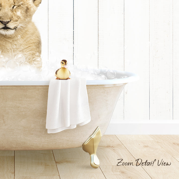 A plush toy duck sits in a bathtub filled with bubbles, with a towel draped over the side and a wooden floor visible in the background.