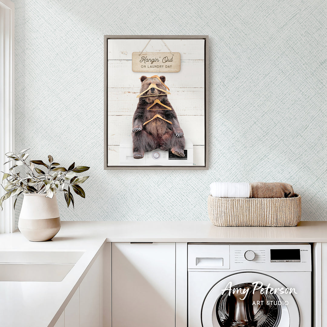 a framed picture of a bear hanging on a wall above a laundry basket, with a potted plant and a washing machine visible in the foreground.