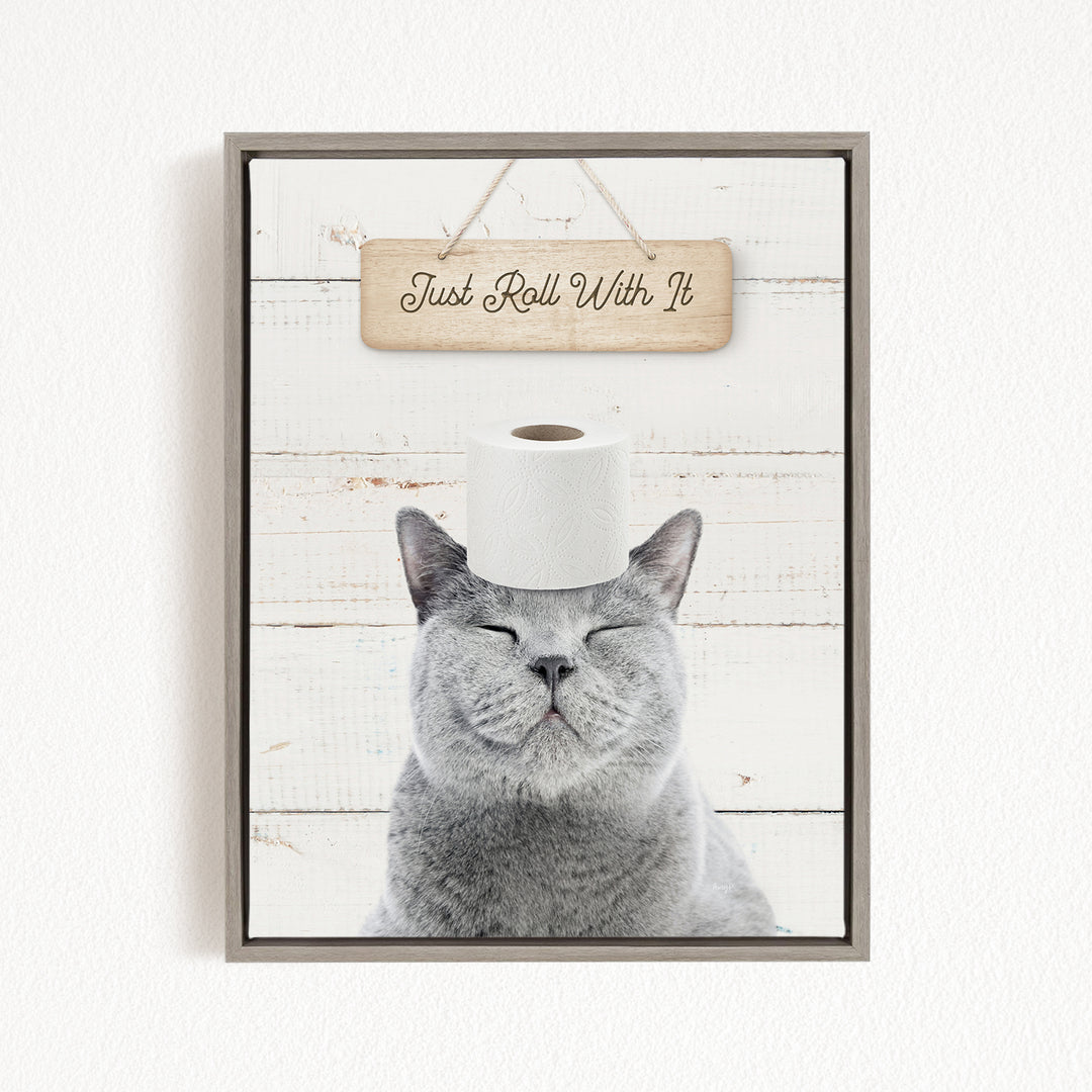 A gray cat with its eyes closed and a roll of toilet paper placed on its head, with a wooden sign above that says "Just Roll With It".