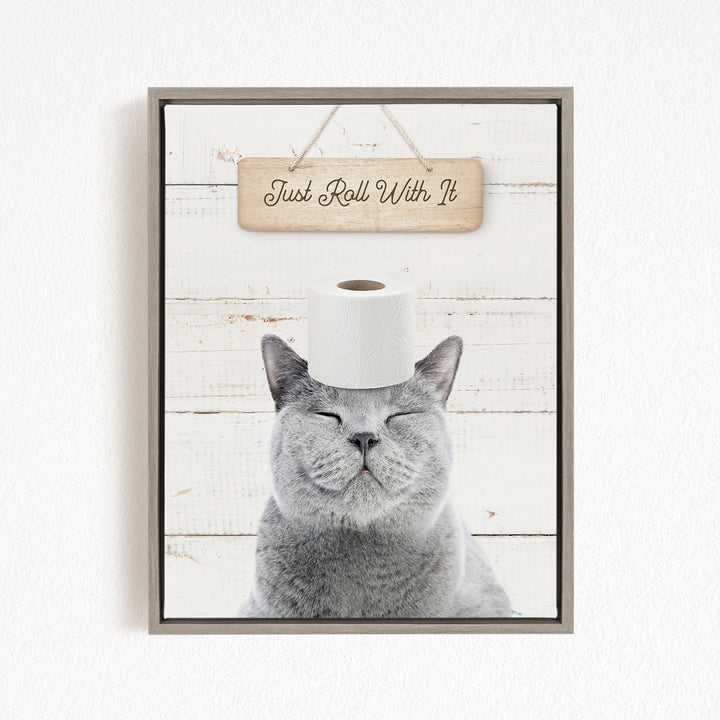 A gray cat with its eyes closed and a roll of toilet paper placed on its head, with a wooden sign above that says "Just Roll With It".