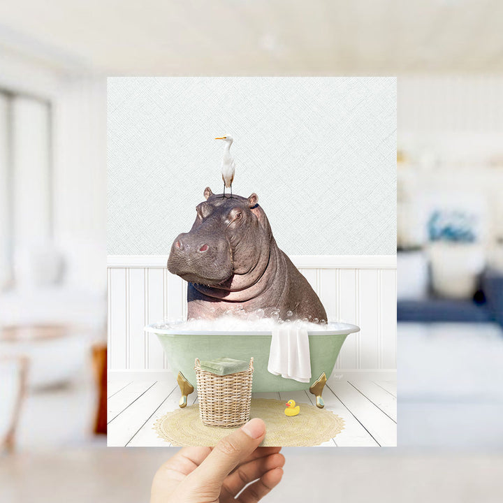 A hand holds up a framed image depicting a hippopotamus sitting in a bathtub, with a bird perched on its head.