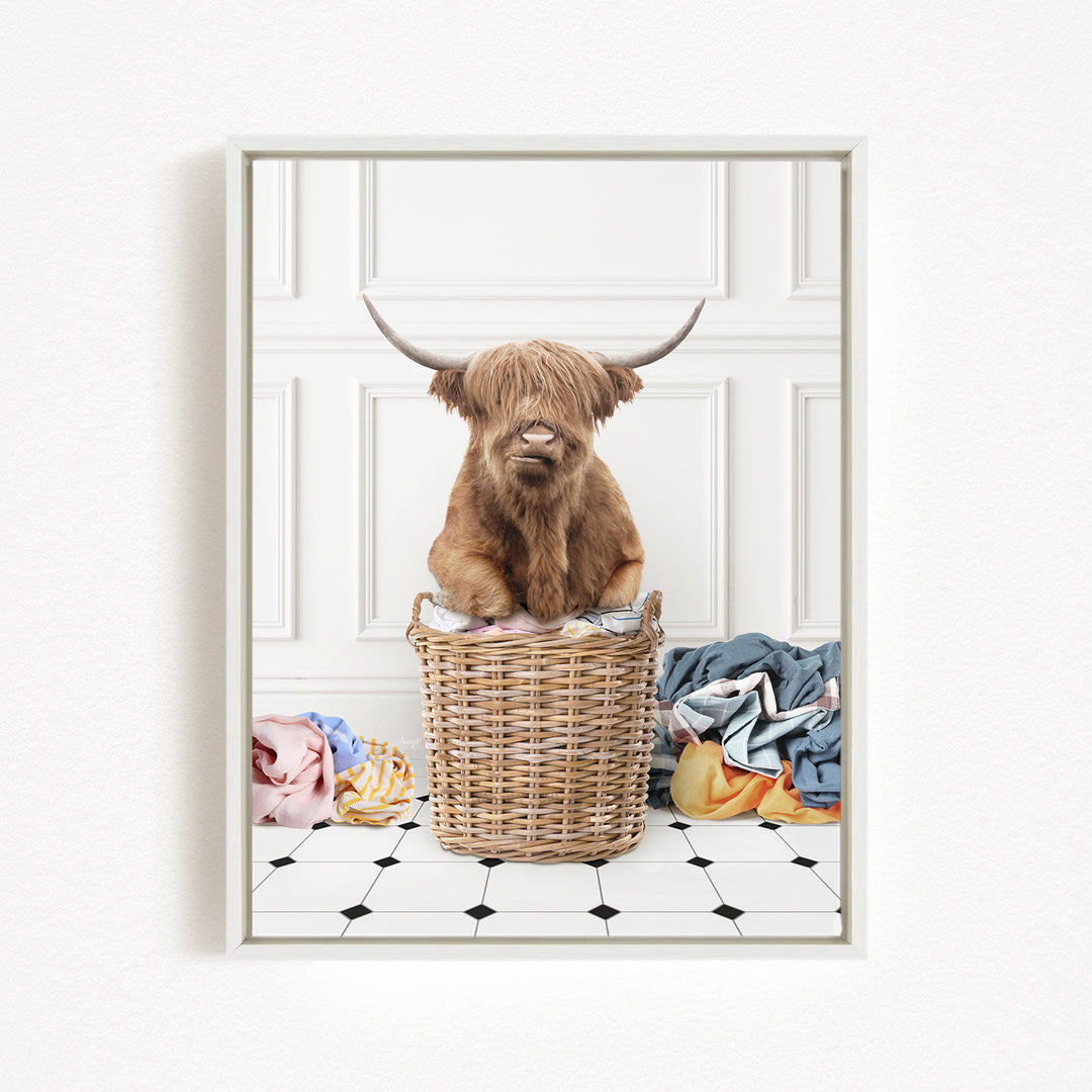 A Highland cow sits in a wicker basket filled with laundry, surrounded by various colorful clothes.