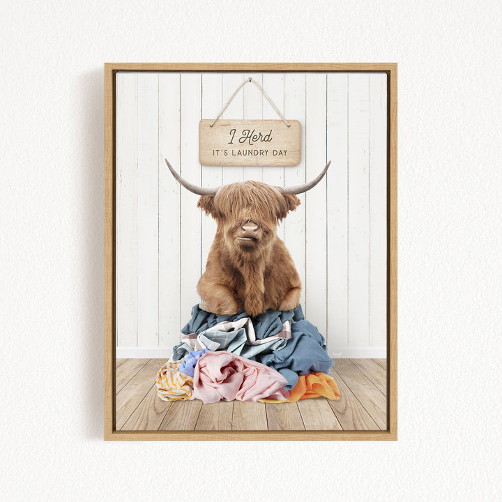 A framed image of a Highland cow with large horns sitting on a pile of laundry, with a sign above that says "I herd it's laundry day".
