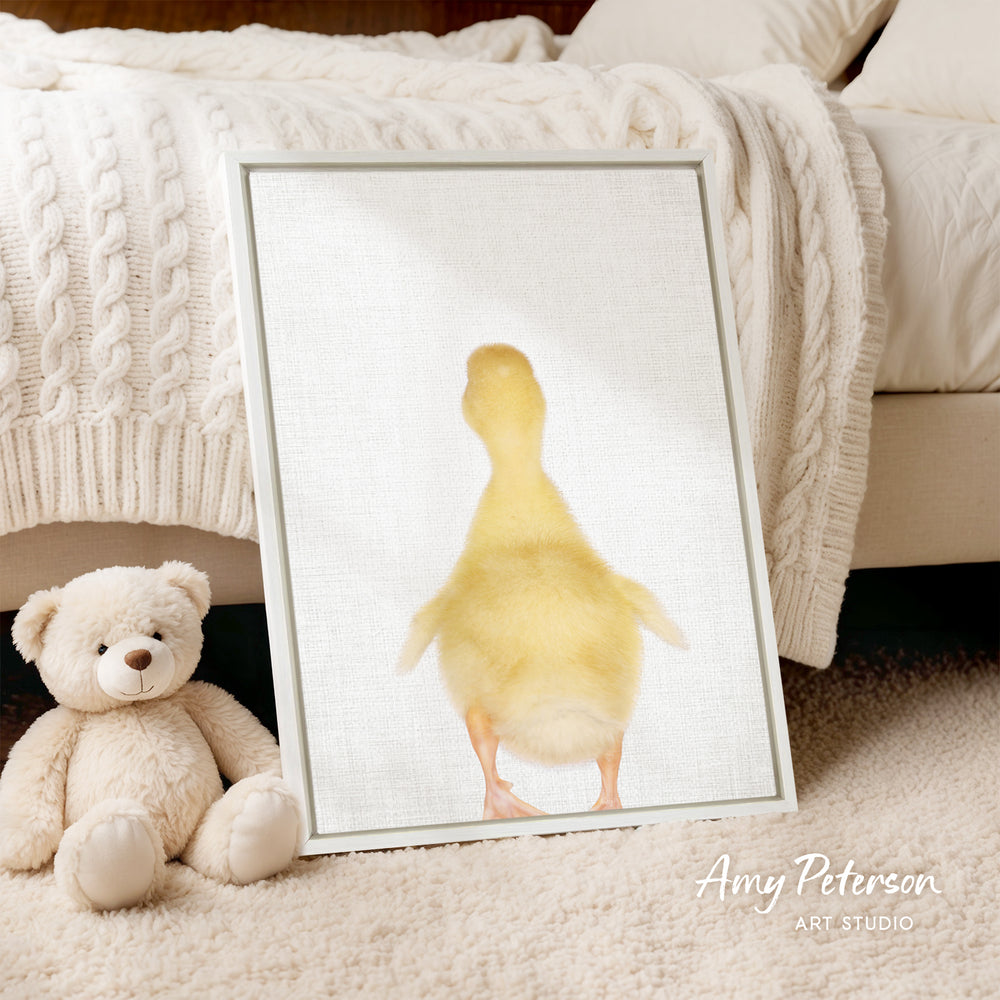 A framed image of a yellow duck is displayed on a white surface, with a white teddy bear sitting next to it.