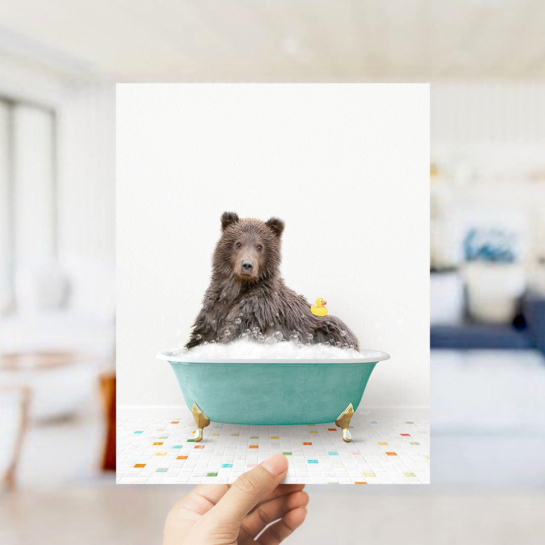 A hand holds up a framed image of a brown bear sitting in a bathtub filled with bubbles, with a yellow rubber duck floating nearby.