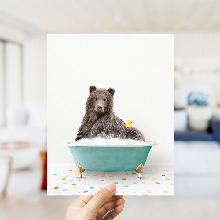 A hand holds up a framed image of a brown bear sitting in a bathtub filled with bubbles, with a yellow rubber duck floating nearby.