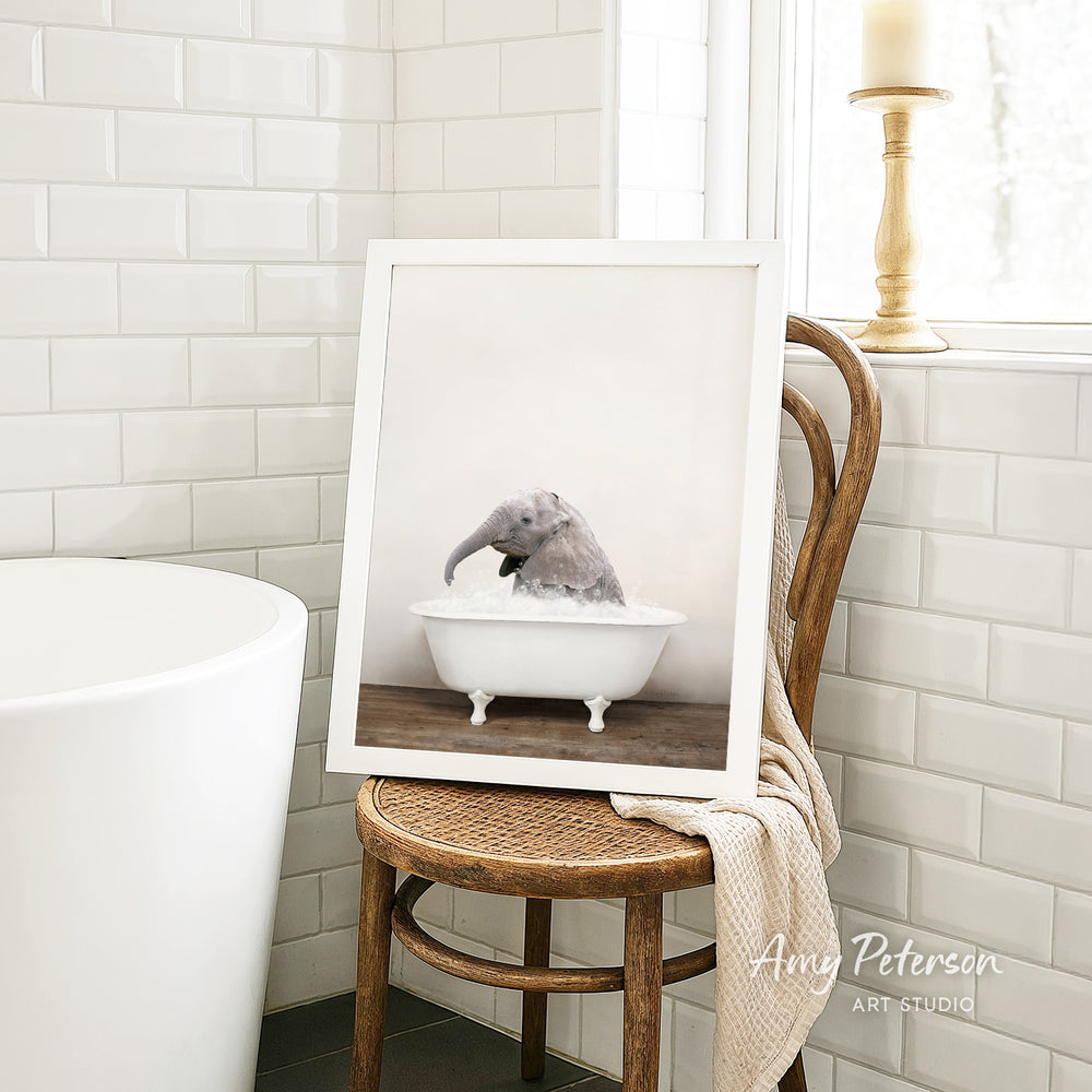 A framed photograph of a gray elephant sitting in a white bathtub, with a towel draped over the back of a wooden chair in a bathroom setting.