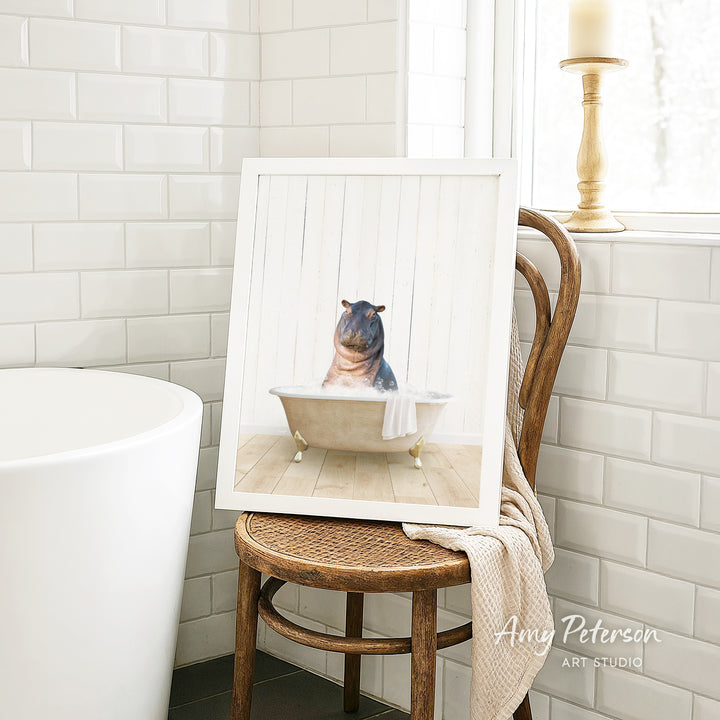 A framed image of a hippopotamus sitting in a bathtub is displayed on a wooden chair in a bathroom setting.