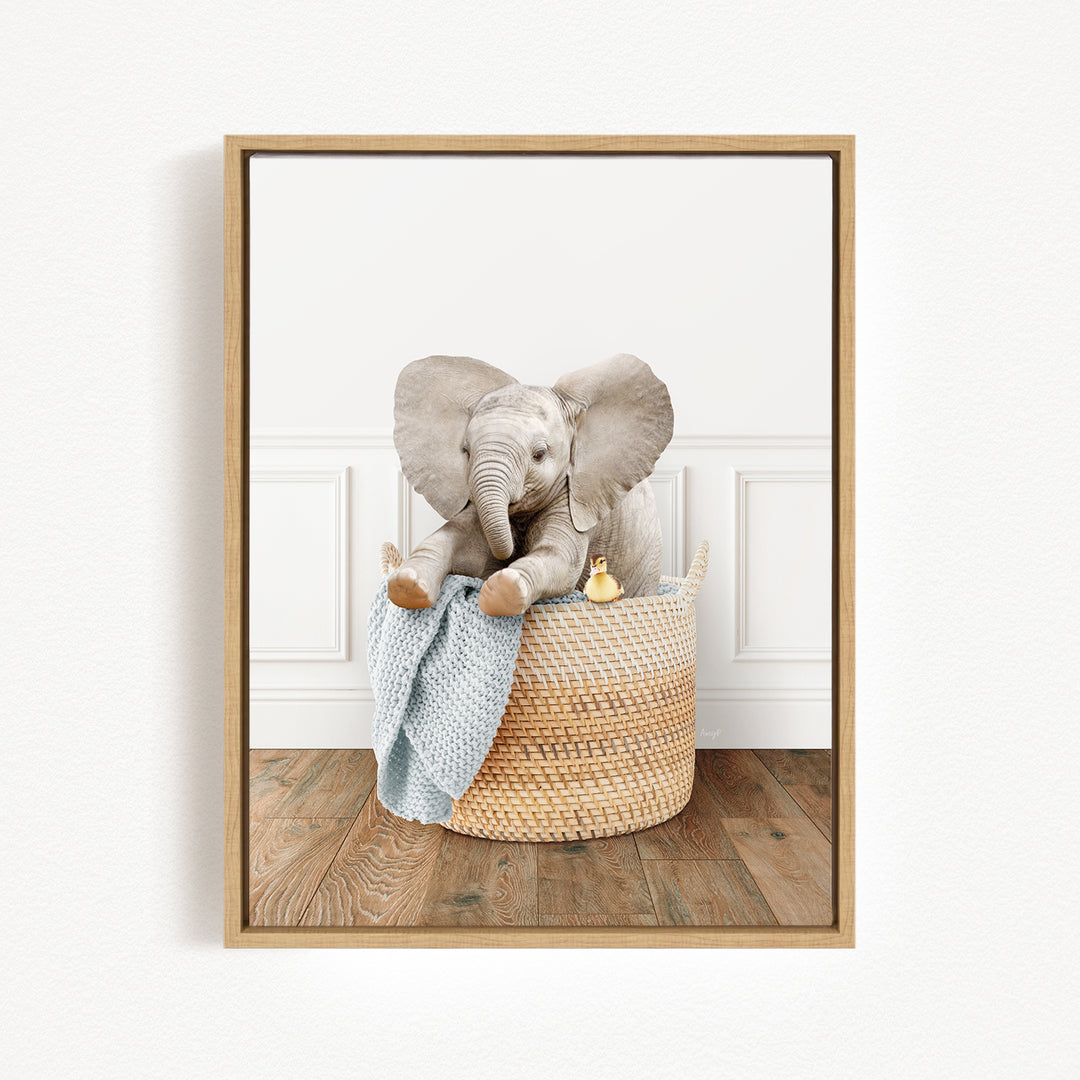 A baby elephant sitting in a woven basket, holding a blue blanket and accompanied by a small yellow toy.