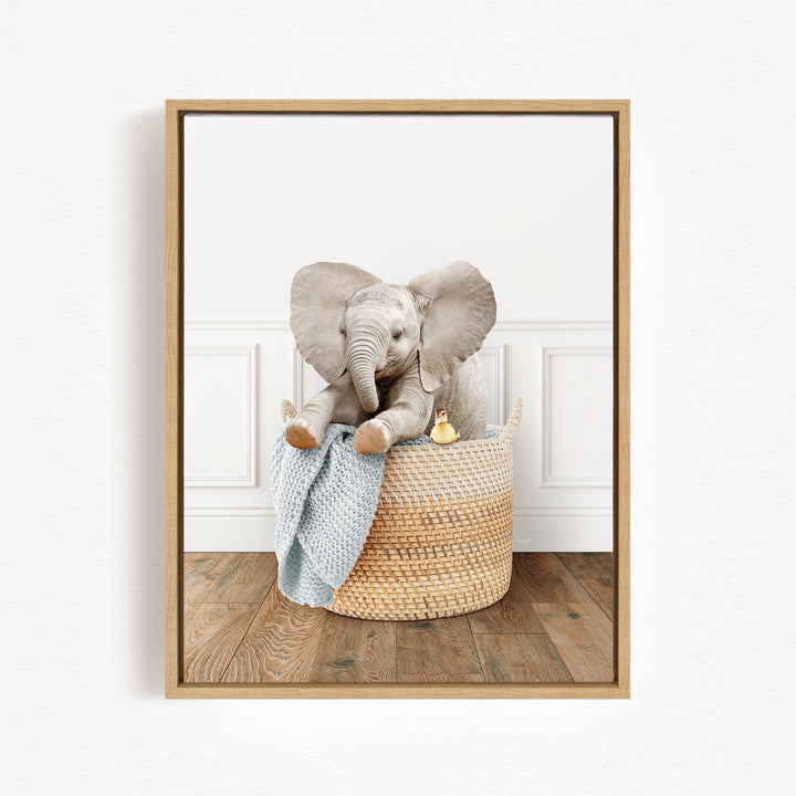 A baby elephant sitting in a woven basket, holding a blue blanket and accompanied by a small yellow toy.
